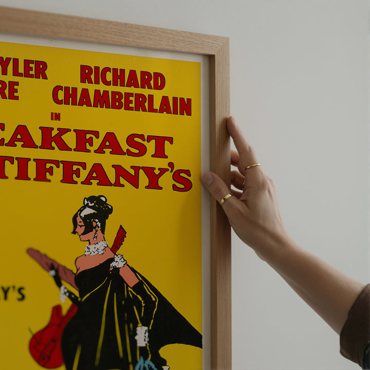Hand holding a wooden frame of a Breakfast at Tiffany's Musical Poster with bright yellow background and red text wall art print